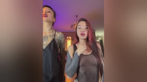 Video of two women, one with a tattooed arm, wearing sheer black tops, in a dimly lit room.