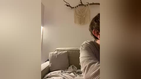 Video of a woman with short brown hair, wearing a textured gray cardigan, sitting on a beige couch with a blanket, in a dimly lit room with a beige wall and macram\u00e9 wall hanging.