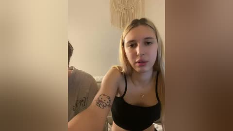 A video of a young woman with long blonde hair, fair skin, and a black tank top, sitting indoors, holding a framed picture.