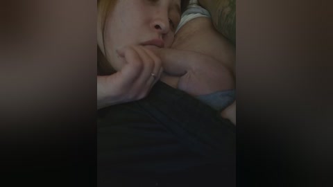 A close-up video of a woman with fair skin and red hair, resting her head on her arm, wearing a gray shirt, in a dimly lit room.