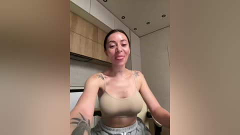 A video of a fit, light-skinned woman with brown hair, wearing a beige sports bra and gray sweatpants, sitting in a modern kitchen with wooden cabinets and recessed lighting.