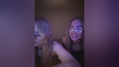 Video of two smiling young women, one with glasses, looking through a doorway, lit by dim purple lighting.
