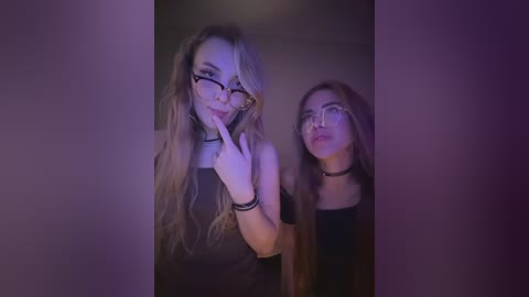 Video of two young women, one with long blonde hair, the other with brown hair, wearing glasses and chokers, posing provocatively in dim lighting.