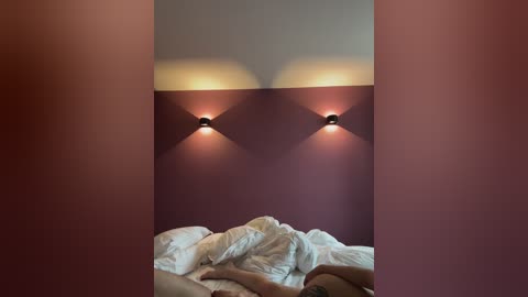 Video of a cozy bedroom with a muted palette. The bed is covered with white sheets, and two wall-mounted sconces emit soft light. The walls are painted in a deep maroon color, creating a warm and inviting atmosphere.