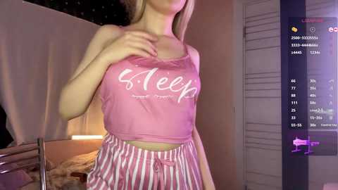 Video of a fair-skinned, blonde woman in a pink, see-through \"Sleep tight, don't let the bedbugs bite\" tank top and striped pajama pants, standing indoors with a dimly lit background.