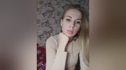 A video of a blonde woman with fair skin, wearing a beige top, sitting in a room with floral wallpaper. She has a thoughtful expression, resting her chin on her hand.