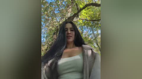 Video of a young woman with long black hair and medium skin tone, wearing a white tank top and light jacket, standing in a sunlit forest with green leaves and blue sky.