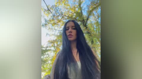 A video of a woman with long, dark hair, wearing a grey jacket, standing in a forest with green leaves and blue sky background.