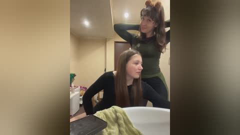 Video of two women in a bathroom. One, with long brown hair, wears a black top, leaning on a sink. The other, with brown hair, wears a green top, playfully touching her hair.