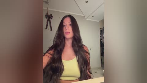 A video of a woman with long, wavy, dark brown hair, wearing a lime-green crop top, standing in a modern, minimalist bathroom with beige walls and a tiled floor.