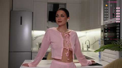 A video of a slender, fair-skinned woman with long dark hair, wearing a pink lace-up crop top and matching skirt, posing in a modern kitchen. The background features white cabinets, stainless steel appliances, and a sleek, minimalist design.