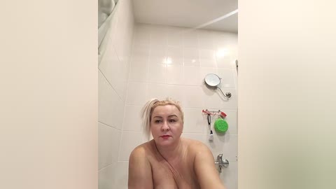 Video of a topless, middle-aged woman with fair skin and blonde hair in a shower with white tiles, holding a red towel.
