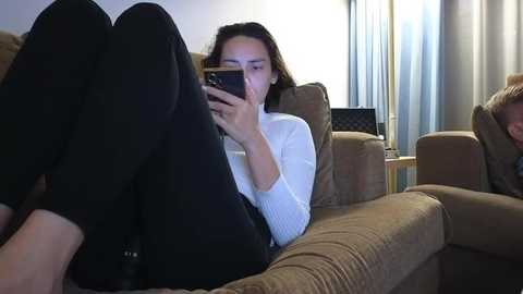 Video of a woman with shoulder-length brown hair, wearing a white sweater and black leggings, lying on a brown couch, engrossed in her smartphone.