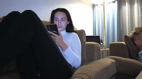 A video of a woman with long black hair, wearing a white sweater and black pants, sitting on a beige sofa, texting, with another person in the background, dimly lit room, beige curtains, and a lamp.
