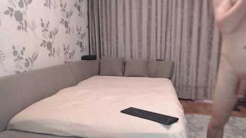 A video of a modern, minimalist bedroom with beige carpet, gray sofa, white floral wallpaper, beige bedspread, and a keyboard on the floor.