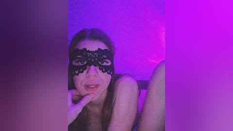 Video of a fair-skinned woman with long brown hair, wearing a black lace mask and a seductive expression, against a purple and pink-lit background.