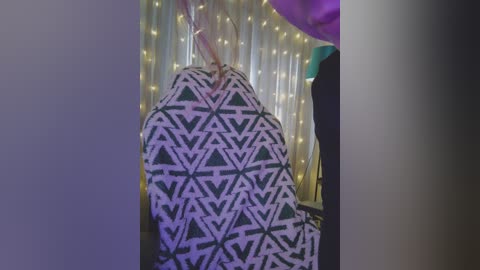 Video of a person with light brown hair wearing a geometric-patterned black and white blanket, sitting in a dimly lit room with string lights and purple fabric hanging in the background.