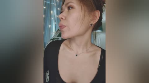 A video of a fair-skinned woman with auburn hair in a black top, adorned with a small necklace and earrings. She gazes thoughtfully to the left, surrounded by softly lit, blurry curtains and greenery in the background.