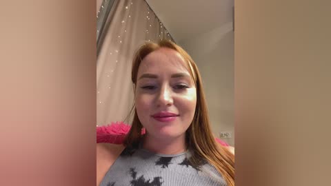 Video of a fair-skinned, red-haired woman with long hair, wearing a tie-dye tank top, standing in a room with a beige curtain and pink fuzzy throw.