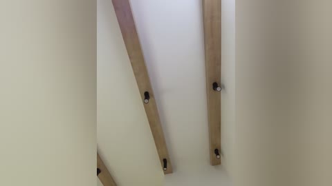 A video of a minimalist ceiling with three wooden beams intersecting at various angles, secured by black metal brackets, against a smooth, white wall.