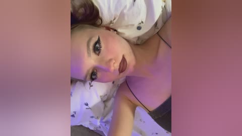 Video of a fair-skinned woman with dark makeup, lying on a bed with a white pillow. She has dark hair and is wearing a black bra, her lips are painted dark red.