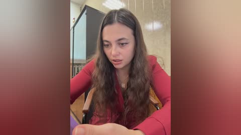 Video of a young woman with long, wavy brown hair in a red blazer, looking thoughtful, seated in a classroom with beige tiled walls and wooden furniture.
