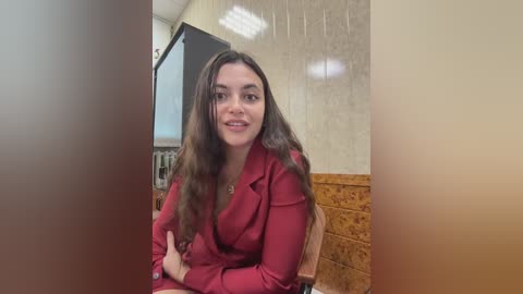 Video of a smiling woman with long brown hair, wearing a red blazer, sitting on a wooden chair with a beige wall and brown laminate flooring in the background.