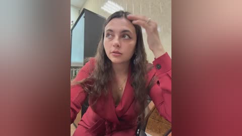 Video of a young woman with long brown hair, wearing a red blazer, sitting in a chair, looking thoughtful, indoors with a brick wall background.