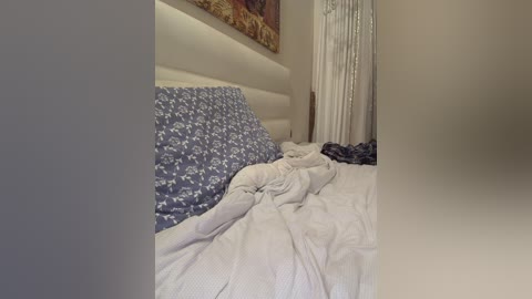 A video of a messy bedroom with a white tufted headboard, a blue floral duvet, and rumpled white sheets. A partly visible painting and sheer curtains are in the background.