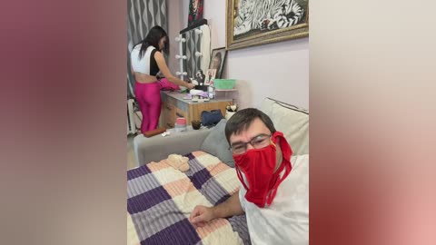 A video of a man in a red mask lying on a bed, a woman in pink leggings working at a desk in the background, a plaid blanket covering the man, and a framed painting on the wall.