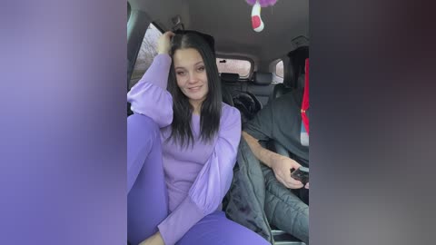 Video of a young woman with long black hair and fair skin, dressed in lavender pajamas, sitting in the backseat of a car, smiling.