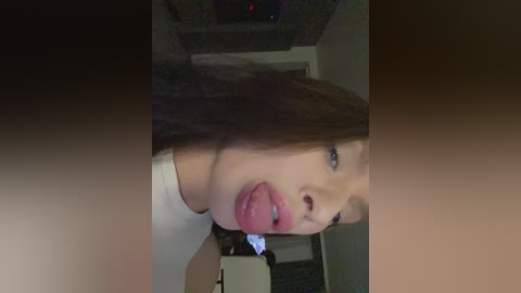 Video of a young Asian woman with long brown hair, fair skin, and light pink lipstick, sticking out her tongue. She wears a white top. The background shows a dimly lit room with a TV and furniture.