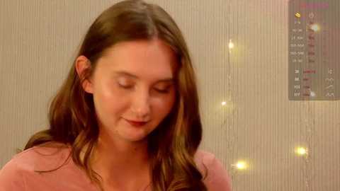 A video of a young woman with long, straight brown hair, wearing a pink top, smiling softly, against a textured beige wall with a calendar and glowing lights in the background.