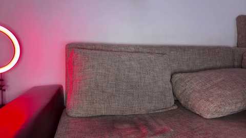 Video of a modern living room featuring a beige, textured fabric sofa with a large circular, red lightbulb on the left, casting a warm glow. The background wall is plain white, creating a minimalist aesthetic.