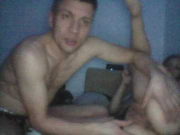 A blurry video of two shirtless men, one with a muscular build and the other leaner, engaged in a sexual act on a bed. The background is dimly lit with indistinct objects, suggesting a private, intimate setting.