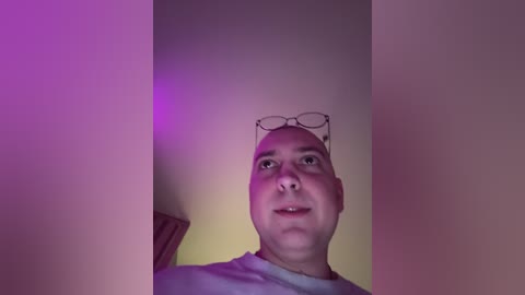 Video of a bald, middle-aged man with glasses, wearing a white T-shirt, standing in a dimly lit room with purple and yellow lighting.