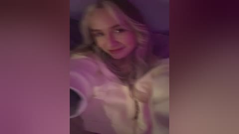 A blurred video of a smiling blonde woman with wavy hair, wearing a white shirt, captured in a dimly lit room with purple and pink hues.