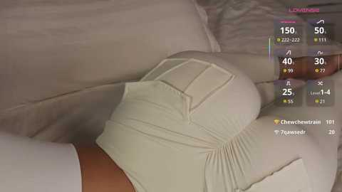 A video of a person lying on a bed with a beige blanket, wearing a beige dress with pleated details. The room is dimly lit, and a digital interface with character statistics is overlaid in the top right corner.