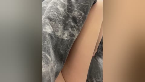 Video of a person's nude, pale-skinned thigh and upper leg, draped in a soft, gray, marbled blanket. The background is plain and neutral, likely a wall, emphasizing the texture and contrast between the smooth skin and the plush blanket.