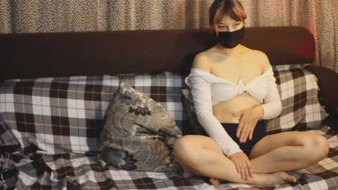 Video of an East Asian woman with light skin, sitting cross-legged on a bed with plaid bedding, wearing a white off-shoulder top, black mask, and black shorts, surrounded by a cozy, dimly lit room.