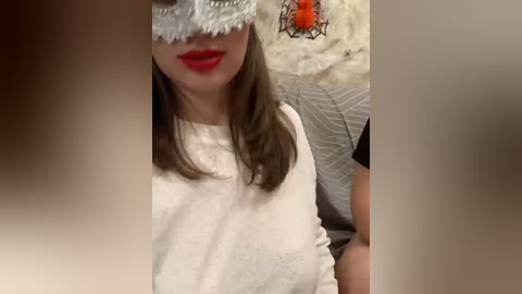 Video of a woman with light skin, brown hair, and red lipstick, wearing a white sweater and a lace mask, sitting on a gray couch with a decorative orange and black candle nearby.