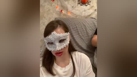 Video of a young woman with fair skin, wearing a delicate, lace masquerade mask, red lipstick, and a white blouse. She is seated on a gray cushioned chair, surrounded by festive, Halloween-themed decorations with orange lights and a spider web.