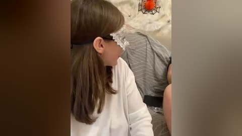 Video of a woman with long brown hair and a white lace headband, wearing a white blouse, sitting in a dimly lit room with a grey-patterned dress in the background.