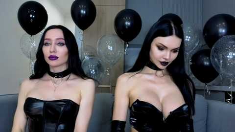 Video of two women with pale skin, long black hair, and dark makeup. They wear black latex dresses, chokers, and black balloons in a modern, minimalist room.