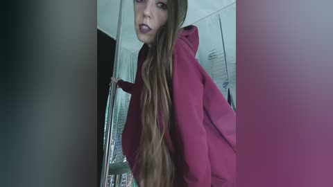 A video of a young woman with long, wavy brown hair, wearing a red hoodie, peeking through a glass door in a dimly lit room, with blurred background.