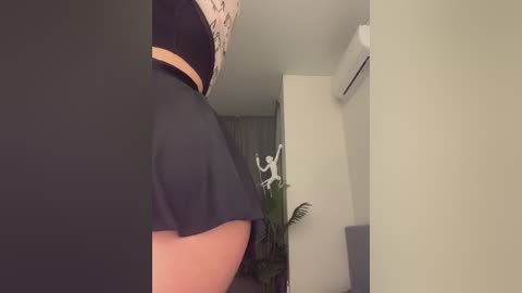 Video of a woman's lower back and buttocks in a black skirt, viewed from the side. Background includes a white wall, a green plant, and an air conditioner.