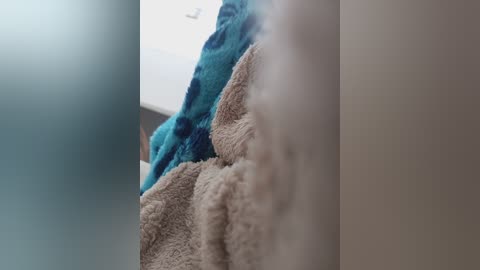 A close-up video of a beige, fluffy towel next to a blue towel with a dark blue pattern, both slightly out of focus. The background is a blurred, light-colored wall.