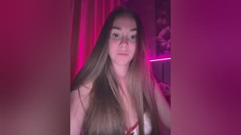 Video of a young, fair-skinned woman with long, straight brown hair, wearing a revealing white lace bra. She's indoors, bathed in pink and purple lighting, with floral wallpaper in the background.