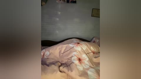 Video of a messy bed with a beige comforter featuring large pink cherry blossoms. The room's walls are plain white, and a small framed picture hangs on the right.
