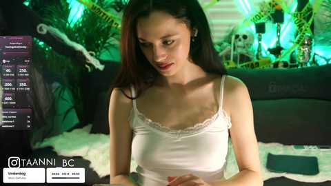 A video of a slender woman with fair skin and long dark hair, wearing a white camisole, seated in a dimly lit, Halloween-themed room with skeletons, green lights, and a dark couch.